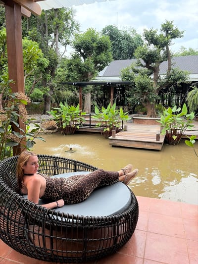A woman is lying on a circular wicker lounge chair, facing to her left. She is wearing a leopard-print one-piece swimsuit and tan sneakers. Her hair is blonde and pulled back. The chair is placed on a reddish-brown tiled patio. Behind the chair is a muddy pond with a small fountain in the center. There are wooden decks extending into the pond, surrounded by lush green vegetation. In the background, there are trees and a building with a dark gray roof. The sky is overcast. The location is a resort in Mae Rim, Thailand.