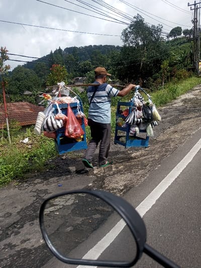 A vendor carries two blue, shelved containers filled with bagged goods on a bamboo pole across his shoulders. He wears a striped shirt, dark pants, a hat, and sandals. He walks along a road with a white line marking the lane. Lush greenery, trees, and hills form the background. Electrical wires stretch across the sky. A blurred motorcycle mirror is visible in the foreground, suggesting the photo was taken from a moving vehicle.