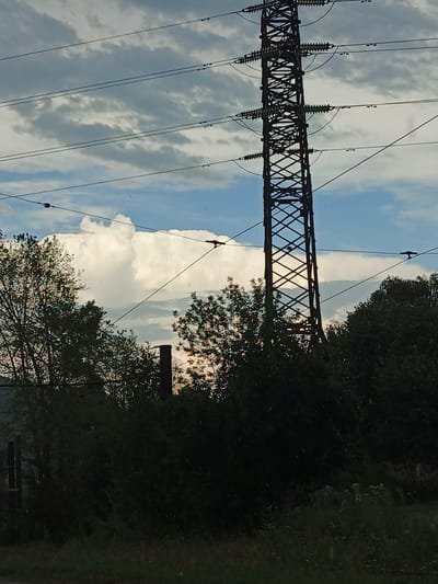 A tall, lattice-like transmission tower dominates the right side of the frame, reaching towards a sky filled with a mix of grey clouds and patches of blue. Several power lines stretch horizontally across the image, attached to insulators on the tower and disappearing into the left side of the frame. In the foreground, dense, dark green foliage of trees and bushes obscures the lower part of the scene, with a dark, vertical structure, possibly a smokestack, visible just to the left of the transmission tower. The overall lighting suggests either early morning or late afternoon, with a soft, diffused light. There are visible streaks of rain or water droplets, indicating current precipitation. The scene appears to be an outdoor, possibly industrial or semi-rural setting, given the presence of the power lines and tower. There is no visible text in the image.