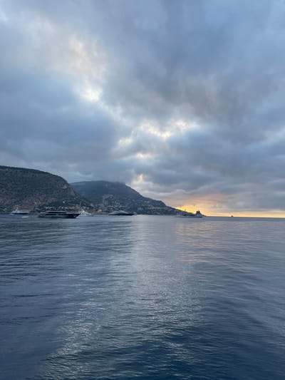A panoramic view captures the azure waters of the Mediterranean Sea off the coast of Èze, France, under a dramatic cloudy sky. Several large yachts are anchored offshore, silhouetted against the moody backdrop. The mountainous coastline of Èze, dotted with buildings, rises steeply from the water, shrouded in mist and clouds. The sun attempts to break through the overcast sky, casting a soft, golden glow on the horizon, hinting at either sunrise or sunset. The water's surface is rippled, reflecting the diffused light from the sky.