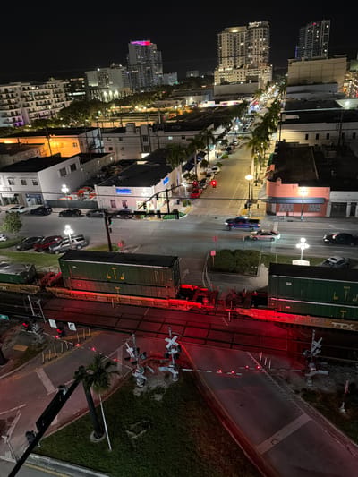 This is an aerial, nighttime photograph taken in Hollywood, Florida, USA. The scene captures a street with car traffic and a railroad crossing in the foreground. Several multi-story buildings, some illuminated and appearing to be apartments or hotels, are visible in the background, along with city lights and palm trees lining the street.

In the foreground, a train with several green and brown freight cars is stopped at a railroad crossing. The crossing gates are down, and red lights are flashing, indicating an approaching or present train. Cars are stopped on the street on either side of the tracks, waiting for the train to pass. The street itself is illuminated by streetlights and the lights from nearby buildings.

The buildings in the background vary in height and architectural style. Some have illuminated windows, suggesting occupancy. The overall impression is of a lively urban environment at night. The photograph is taken from a high vantage point, possibly from a taller building overlooking the street and railroad.