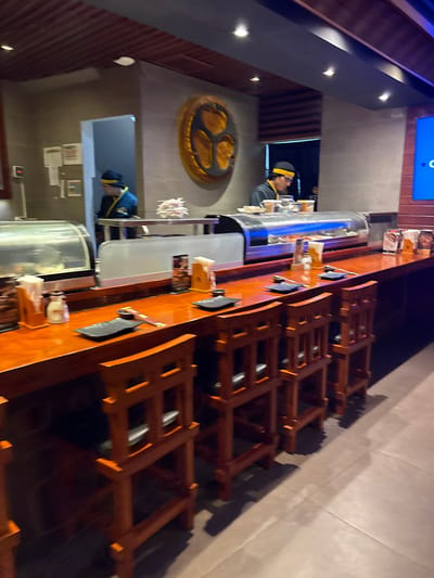 The image depicts the interior of a sushi restaurant in Magdalena del Mar, Peru. A wooden bar with a polished, orange-toned surface runs horizontally across the frame. Several wooden bar stools with an open, ladder-back design are positioned in front of the bar. On the bar, various items are visible including black rectangular plates, chopsticks, small white containers, a salt shaker, and paper napkins. Behind the bar, a refrigerated display case filled with sushi is prominent. Two individuals, presumably chefs or staff, are seen working behind the bar. One is wearing a blue uniform and a yellow headband, and is partially visible through a doorway. The other, also in a blue uniform and yellow headband, is more clearly visible, interacting with the sushi display. The background features a grey tiled wall with a large, circular, golden decorative piece. The lighting is dim, with spotlights illuminating the bar area.