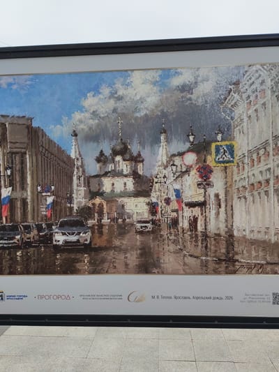 The image is a painting depicting a street scene in Yaroslavl, Russia, during what appears to be a rainy day. The artwork captures a view of a street with a prominent church with multiple domes on the right side. Buildings line both sides of the street, with the one on the left appearing to be a more modern, multi-story structure. Several cars are parked or driving on the wet street, reflecting the overcast sky and surrounding lights. Russian flags are visible, hanging from poles. The painting is framed and displayed outdoors, as suggested by the background which includes a portion of a ceiling or overhang and the ground. The bottom of the image contains text and logos. One logo reads "МЭРИЯ ГОРОДА ЯРОСЛАВЛЯ" (Mayor's Office of the City of Yaroslavl). Another text line reads "М. В. Теплов. Ярославль. Апрельский дождь. 2026" (M. V. Teplov. Yaroslavl. April Rain. 2026), indicating the artist, location, title of the artwork, and year of creation. Further text provides exhibition hall details, including an address and phone number.
