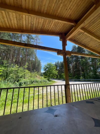 The image depicts an outdoor wooden structure with a slatted ceiling and exposed wooden beams, likely a pavilion or gazebo. Through an opening in the structure, a view of a forest with tall pine trees is visible under a bright blue sky with sparse white clouds. A paved road or pathway is seen in the middle distance, winding through the trees. In the foreground, a black metal railing separates the structure from a grassy area. The ground beneath the railing is covered in green grass and some low-lying plants. The front of the image shows a portion of a patterned surface, possibly a table, made of a light-colored material with dark gray geometric designs. The scene appears to be a tranquil natural setting, possibly a rest stop or recreational area, located in the vicinity of Valkosel, Bulgaria. No people are visible in the image.