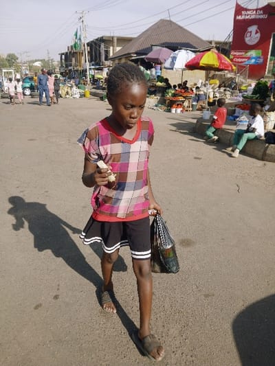 This image depicts a daytime street scene in Kwekwe, Zimbabwe, characterized by market activity.
The primary subject is a young, dark-skinned female, approximately 8-12 years old, walking forward on an asphalt road. She wears a multi-colored plaid short-sleeved top over a red undershirt, black shorts with two white horizontal stripes on the lower hem, and dark open-toed sandals. Her hair is styled in short braids. In her right hand, she holds a light-colored, elongated object near her mouth. In her left hand, she carries a dark bag with a light, geometric pattern. Her gaze is directed downwards and slightly to her right. A distinct, elongated shadow of her figure is cast on the road to her left, indicating sunlight from the right.
In the midground to the right, a vibrant market operates. Two children are seated on a low concrete barrier: one in a red shirt and green pants, the other in a white shirt and light green pants. Produce stalls, displaying items such as red tomatoes, are visible under a red and yellow striped umbrella and a blue and white umbrella. Behind these stalls, a multi-story building features a large red billboard with "YANGA DA MURMUSHINKU" and "Colgate" branding. Another building with a brown, peaked roof is situated behind the market stalls.
To the left of the central figure, further down the road, other individuals are visible, including a man in a blue shirt. A light green vehicle is parked or positioned near the side of the road. More market activities or street vendors with various objects on the ground are present. Buildings with power lines overhead and green flags on a pole are also discernible in the distant background on the left. The road itself is paved asphalt, showing minor cracks and stains. The sky appears clear and bright.