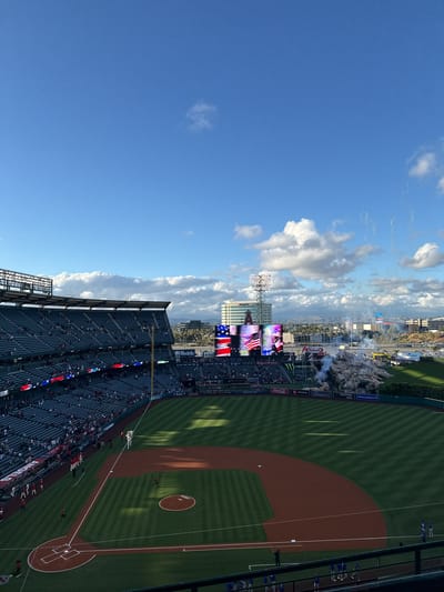 The image is an oblique aerial view of Angel Stadium in Anaheim, California, United States. It's game day with a clear blue sky and scattered clouds. The stadium is filled with spectators. The baseball field is green, and the infield is reddish-brown. The outfield fence is visible, marked with advertising. The scoreboard displays American flags, and advertising is apparent along the outfield fence. In the background, one can observe what appear to be the foothills of the Santa Ana Mountains. There are several light poles, a video screen, and a mountain-like structure in center field. The stadium is located in Anaheim, as evidenced by the cityscape in the distance.