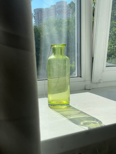 A light green glass bottle with a thin wire handle is placed on a white windowsill in front of a window. The bottle is partially obscured on the left by a dark grey curtain. Through the window, trees and a multi-story building are visible against a bright blue sky. Sunlight casts a long shadow of the bottle onto the windowsill. The scene is set indoors, on what appears to be a sunny day. There are no people or animals present. The environment suggests a residential setting.