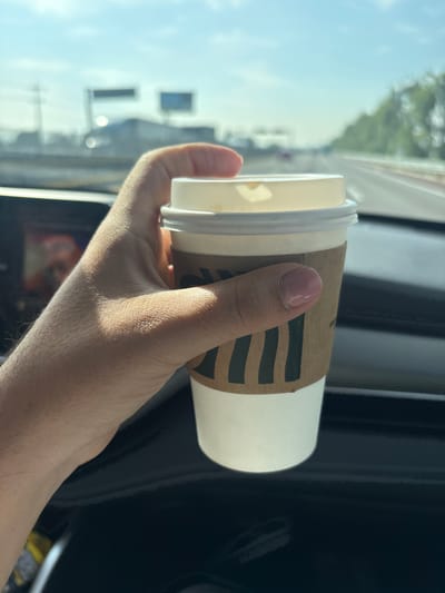 A hand is holding a Starbucks coffee cup with a sleeve in a car. The scene is captured from inside a vehicle, looking outward toward the exterior environment. The background shows a blurred view of what appears to be a street or highway, with trees, buildings, and vehicles visible under a bright sky. The photo was most likely taken in a car and not from San Lucas el Grande, Mexico. The hand holding the coffee cup is light-skinned, and the fingernails appear to have pink nail polish. The coffee cup is white with a brown Starbucks sleeve, and a white lid. The interior of the car is visible, with dark panels and a glimpse of the dashboard.