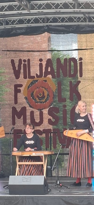 In the city of Viljandi, Estonia, a live outdoor musical performance is taking place on a stage. Two women are the focal points of the image. The woman on the left, seated, is playing a stringed instrument resembling a dulcimer, placed on a wooden stand. She wears glasses and a black long-sleeved shirt with an embroidered circular design. The woman on the right, standing, is holding a triangular stringed instrument and looking to her right. She wears a black long-sleeved shirt with a similar embroidered design and a long, vertically striped skirt in various colors. A banner behind them displays the text "VILJANDI FOLK MUSIC FESTIVAL" in large, stylized letters, with a circular graphic in the center. Stage lighting trusses and a black tarp are visible above the banner, and a large black speaker is positioned in the foreground. Microphones and wires are also present on the stage, indicating a performance in progress. The background appears to be a brick wall and some greenery.