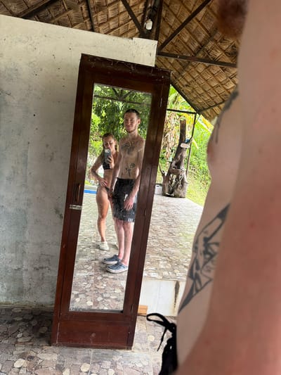 The media file shows a mirror reflecting a young couple. The woman is wearing a black bikini and the man is wearing black shorts. They are both shirtless and have tattoos. They are posing for a selfie in a tropical outdoor setting. The background of the reflection shows lush greenery, suggesting a warm climate. The mirror itself is framed in dark wood and appears to be part of a rustic structure, possibly an open-air changing room or bathroom. The setting has a thatched roof visible in the upper left portion of the frame. The overall mood is casual and relaxed. The floor in the reflection is tiled with a pattern of neutral-colored stones. The original image also captures parts of the environment outside the mirror, including the textured wall of the structure and the thatched roof, as well as a portion of the person holding the camera, whose arm with tattoos is visible in the upper part of the frame. The lighting suggests it is daytime, possibly morning or afternoon.