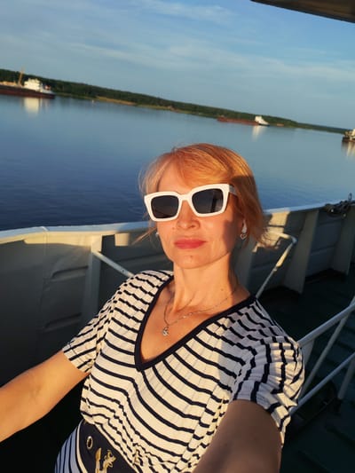 A woman with blonde hair, wearing white-rimmed sunglasses, a striped V-neck shirt, and a necklace, is taking a selfie on a boat. She is positioned in the foreground, with the water and a tree-lined shore in the background. Two cargo ships are visible in the distance. The scene is illuminated by the warm light of a sunset. The location is identified as the city of Shchetinino, Russia.