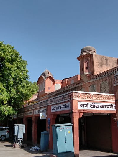 The image shows a commercial building in Jaipur, India. The building's facade is made of red stone, with ornate architectural details including arches, domes, and carved patterns. Signage in Hindi indicates that several businesses are located within the building.  The sign on the top reads "Garg Watch Company", and the sign on the bottom says "Motilal Sontilal Watch Company". The buildings appear to be located on a commercial thoroughfare. A large green tree is positioned to the lower left. In front of the building, on the right, there is a small blue metal electrical box, a metal transformer, a white car with 5 wheels, and sidewalk. The sky is clear and blue.