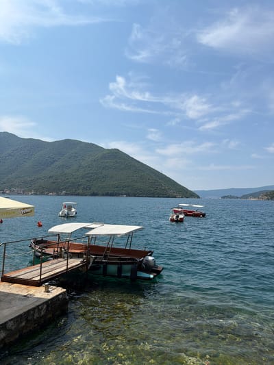 The image depicts a serene view of the Bay of Kotor near Perast, Montenegro. In the foreground, a wooden walkway extends from a stone pier into the clear, turquoise water, where two boats with white canopies are moored. The boats are designed for passengers, with seating and ample shade. The water is calm, with sunlight glinting off its surface, revealing the rocky seabed below.
Further out on the bay, a small white motorboat navigates the water, and in the middle ground, two larger boats, one red and one white, are anchored. The backdrop is dominated by a lush, green, tree-covered mountain that rises majestically from the water's edge. The sky above is a bright blue, dotted with wispy white clouds, indicating a sunny day. The scene is one of tranquility and natural beauty, characteristic of the Montenegrin coast.