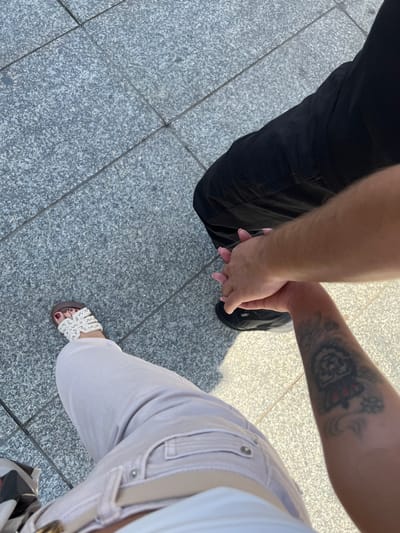 The image shows a close-up, overhead view of two people holding hands while standing on a tiled sidewalk. One person is wearing black pants and black sneakers. The other person is wearing light-colored pants and white sandals with intricate beading. The person wearing the sandals also has a tattoo on their arm, which appears to be of a crown and some floral elements. The sunlight casts shadows on the ground, suggesting it is daytime. The setting appears to be an urban environment, possibly in Katowice, Poland, given the grey, speckled tiles of the sidewalk. The focus is on the hands clasped between the two individuals, implying a moment of connection or togetherness. No text is visible in the image.