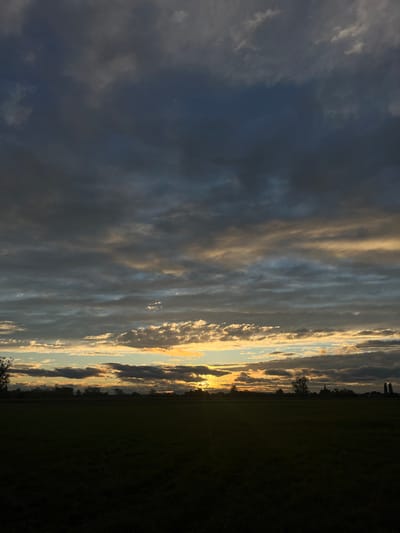 The image depicts a rural landscape at sunset in Hœrdt, France. The scene is dominated by a layered sky, with a band of bright yellow and orange light from the setting sun visible on the horizon, illuminating scattered altocumulus clouds. Above this, the sky transitions to dark grey and blue-grey altostratus clouds, with some darker cumulus formations. The foreground consists of a dark, underexposed expanse of flat agricultural land or grassland, which occupies approximately the lower third of the frame. Along the distant horizon, a dark silhouette of trees is visible, with a prominent church spire and several other buildings or structures from a village visible towards the right side. No individuals or active subjects are present.