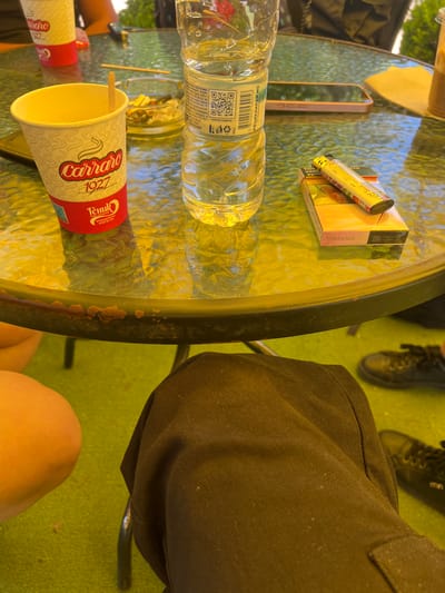 Here is a forensic description of the image:

**Scene:** An outdoor scene, possibly a cafe or patio, in the city of Cherven Bryag, Bulgaria, based on the Carraro cup which is from the local brand Генук.

**Objects:**
*   A round, glass-topped table, showing signs of wear with spots of rust.
*   On the table:
    *   A partly filled water bottle.
    *   A pink cigarette pack labeled "Sobranie."
    *   A lighter.
    *   A smartphone.
    *   A disposable coffee cup.
    *   A cigarette ashtray with cigarette butts.
    *   A second disposable coffee cup.

**People:**
*   Partial views of individuals, including:
    *   Someone wearing black shoes.
    *   Someone wearing black pants.
    *   A bare knee and thigh of a person sitting at the table.

**Environment:**
*   A green surface, possibly grass or an artificial surface, is visible under the table.

**Inferences:**
*   The scene suggests a casual outdoor setting with people relaxing and consuming beverages and cigarettes.