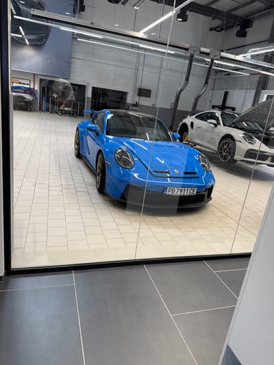 The image depicts a blue Porsche 911 GT3 parked in a brightly lit, modern automotive workshop or showroom. The car is the primary subject, with its sleek design and vibrant color catching the eye. It is positioned on a tiled floor, which appears to be light-colored and laid out in a grid pattern.
In the background, another Porsche, a lighter color, is visible, partially obscured and possibly on a lift, suggesting automotive service or display. Various workshop equipment, such as exhaust extraction hoses and tools on carts, are present, indicating a functional space. The environment is clean and organized, with industrial lighting fixtures visible on the ceiling.
The scene is viewed through a glass partition, which creates reflections and adds a layer of depth to the image. The foreground shows a darker tiled floor, separated from the workshop area by the glass.
The license plate of the blue Porsche reads "PB 7911 CE", and beneath it, the text "Porsche Zentrum Stuttgart / Flughafen" is visible, indicating that the car is a Porsche from the Stuttgart airport dealership. This suggests the image was taken at a Porsche dealership or service center.
While the image is set within a commercial space, there are no people visible in the frame. The focus is entirely on the vehicles and the pristine environment of the automotive facility. The precise location in Radinovo, Bulgaria, is not explicitly indicated by any visual cues within the image itself; this detail would likely be external context.
