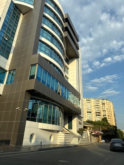 The image shows a street scene in Baku, Azerbaijan. A modern, curved building with many windows dominates the left side of the frame. The building has dark gray panels and white accents, with security cameras mounted on its facade. To the right of this building, a tall, older apartment building with numerous balconies and windows is visible, typical of Soviet-era architecture. A dark-colored car with a license plate starting with "90-YB" is parked on the right side of the street, facing away from the camera. The sky is blue with scattered white clouds. The scene appears to be set during the daytime, with the sun casting shadows. The street itself is paved, and there is a sidewalk with a low chain barrier.