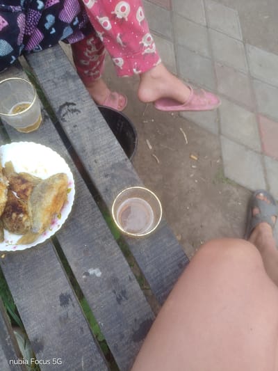 The image depicts a casual outdoor scene, likely in a backyard or patio area. Two individuals are partially visible, seated on a weathered wooden bench.
On the left, a person wearing patterned pajama bottoms with pink reindeer motifs and a dark, patterned robe is seated. Their feet are bare, and one is resting on a pink slipper. In front of them, on the bench, is a plate of fried fish and a glass of light-colored liquid, possibly beer.
To the right, another person is seated, their bare leg and knee prominently featured in the foreground. They are wearing a grey sandal on their visible foot. On the bench, near the plate of fish, is another glass containing a darker liquid, possibly tea or coffee.
The ground adjacent to the bench is paved with diamond-shaped tiles and a dirt path with scattered cigarette butts. The overall impression is one of a relaxed, informal meal or gathering outdoors. There is no specific indicator of the location within Sestrynivka, Ukraine, beyond the general outdoor setting.