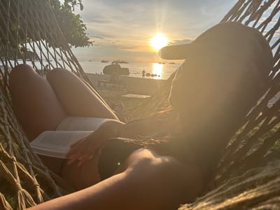 The image displays a human individual relaxing in a woven rope hammock on a beach in Koh Tao, Thailand, during sunset. The person is positioned in the foreground, facing away from the camera and towards the setting sun over the water. They are wearing a dark baseball cap, a dark-colored top, and light-colored shorts or underwear, exposing their legs. An open book with white pages is held in their hands, resting on their lap.
The strong, golden-orange sunlight emanates from the sun low on the horizon, creating a distinct silhouette effect on the subject and the foreground hammock, accompanied by noticeable lens flare. In the midground, a sandy beach is visible, populated by several indistinct figures including a child-like individual near the water's edge and another person seated on a wooden bench. Large, dark rock formations are present along the shoreline. The background features a calm body of water with multiple cargo or passenger vessels visible in the distance. The sky above the horizon displays a gradient from intense orange around the sun to lighter blue-gray, with scattered clouds. Green foliage is visible in the upper left background, indicating adjacent land.