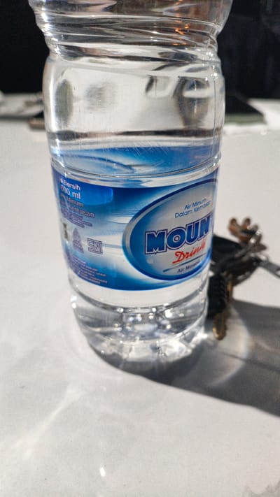 A close-up, slightly angled shot shows a clear plastic bottle of Mountin' Drinking Water, an Indonesian brand, resting on a white, textured surface. The bottle is approximately two-thirds full of water, with subtle ripples visible on its surface. The label, primarily blue and white, features the brand name "MOUNTIN'" in large blue letters within an oval shape, and below it, smaller red text reads "Drink's" and then "Air Mineral". On the left side of the label, smaller text is visible, including "600 ml" and "Air Minum Dalam Kemasan". A small SNI (Standar Nasional Indonesia) logo is also present on the label. To the right of the bottle, out of focus, is a set of keys, possibly attached to a dark lanyard or keychain. The lighting creates soft shadows and highlights on the bottle and the surface it rests on. The background is dark and out of focus, obscuring any specific environmental details. No people or explicit actions are depicted, and the scene appears to be indoors, likely on a table. The image lacks any contextual information regarding the city of Uteungeulinggang, Indonesia.