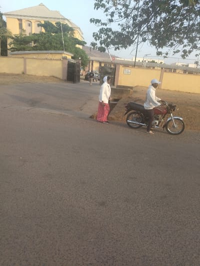 The scene is taken in New Karu, Nigeria, near the coordinates 9.003 latitude and 7.561 longitude. A man is riding a motorcycle on a paved road, and a woman is walking in the opposite direction. The man is wearing a white cap and a grey t-shirt. The woman is wearing a white hijab and a red and white dress. The motorcycle is red and black. The road is paved and there are some trees and bushes on the sides. There is a building in the background. The building appears to be a multi-story structure with a yellow exterior. There is a black gate and a sign that is partially visible. The gate appears to be closed. The motorcycle is stopped at the intersection, facing left. The man appears to be looking forward and the woman appears to be looking to the right.