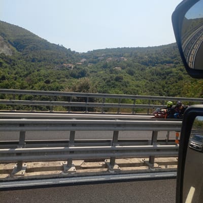 The image captures a view from a vehicle traveling on a highway in Vezzi Portio, Italy. The scene is dominated by a lush, green mountainside covered in dense trees. Scattered among the trees are several houses, suggesting a rural or semi-rural residential area. The sky is clear and blue, indicating a bright, sunny day.
In the foreground, a highway guardrail made of horizontal metal bars runs across the frame, separating the road from the mountainside. On the road, two people are riding a motorcycle. The rider in front wears a bright yellow helmet and a dark jacket, while the passenger behind them wears a patterned shirt and blue jeans, also with a helmet. They are traveling in the same direction as the viewer's vehicle.
To the right of the image, a side-view mirror of the vehicle is visible, reflecting the highway curving ahead and the mountains in the distance. Further down the road, a silver car is visible. The overall impression is one of a scenic drive through the Italian countryside on a clear day. There is no visible text in the image.
