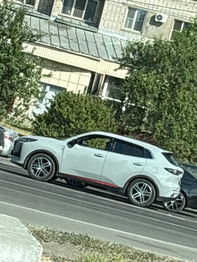 The image captures a light-colored crossover SUV parked on a city street in Biysk, Russia. The vehicle is positioned in what appears to be a lane of traffic, with other vehicles visible nearby, including a dark-colored SUV to its right and a light-colored sedan behind it. The SUV has a red accent stripe running along its lower body and distinctive silver alloy wheels. In the background, a multi-story building with a brick facade and numerous windows is visible, partially obscured by lush green trees. An air conditioning unit is mounted on the exterior wall of the building. The scene is set during daylight, suggested by the lighting and the shadows cast. The overall environment indicates an urban setting, likely a residential or commercial area.