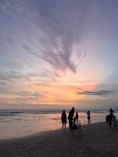 This image depicts a beach scene at sunset in Kerobokan Kelod, Indonesia. The sky displays a gradient from vibrant orange and pink at the horizon to purple and blue in the upper quadrants, with scattered cirrus and stratocumulus clouds. A distinct, diagonal shadow line is visible across the upper right portion of the sky. The ocean features small waves breaking near the shore, reflecting the sunset colors. The dark sandy beach, wet in the foreground and near the water, shows visible footprints and minor debris. Eight human figures are present, silhouetted against the bright sky. In the left-center, an adult male bends towards a child or small object, standing near an adult female. A light-colored dog is in the foreground, walking from left to right on the wet sand, carrying an object in its mouth. To the right, an adult female and a young child stand on the sand; the adult appears to be adjusting clothing. Another adult female stands partially submerged in the water in the central background, facing the horizon. An additional adult female stands alone near the water's edge on the far left. All individuals are engaged in standing, walking, or observing the environment.