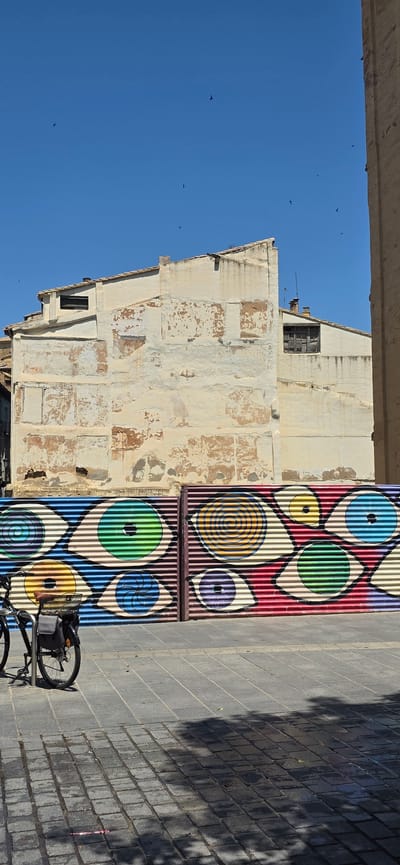 The image depicts a street scene in Huesca, Spain, on a sunny day with a clear blue sky and several birds flying. In the foreground, a bicycle with a bag on the back is parked on a paved sidewalk. Behind the bicycle, a corrugated metal fence is adorned with a vibrant mural of colorful eyes. The mural features various eye designs, some with concentric circles and different colored irises. The fence runs horizontally across the frame, dividing the lower portion of the image from the upper portion.
In the background, an old building with peeling paint and exposed brick is visible. The building has a tiled roof and several windows, one of which appears boarded up. The architectural style suggests an older European structure. The overall scene is a juxtaposition of urban art, everyday life (represented by the bicycle), and aged architecture under a bright sky. The shadows cast on the pavement indicate the time of day and the strong sunlight.