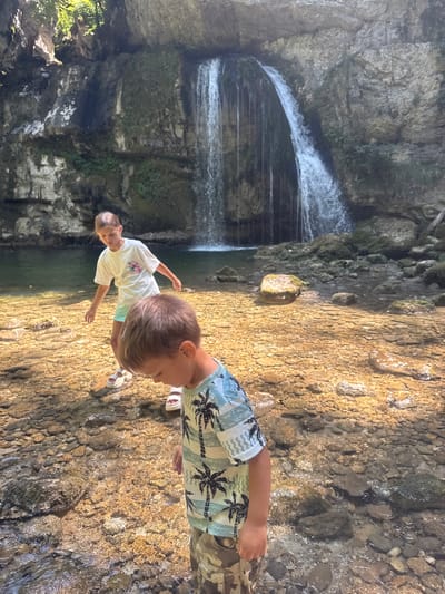 This is a media file of two children by a waterfall. The subjects are a young boy and a young girl. The boy is in the foreground, facing away from the camera and looking down at the water. He is wearing a light-colored, short-sleeved shirt with a palm tree pattern and camouflage shorts. The girl is further back, facing the boy and looking down. She is wearing a white t-shirt with a floral design and light-colored shorts.
The scene is a natural outdoor setting with a waterfall cascading down a rocky cliff face into a pool of water. The ground around the water is made up of small, wet stones and larger rocks. Sunlight is dappling through the trees, suggesting it is daytime, likely late morning or early afternoon. The overall atmosphere appears relaxed and joyful, with the children engaged in exploring the natural environment.
Notable details include the texture of the rocks and the clear water reflecting the sunlight. The presence of the waterfall and rocky terrain indicates a natural, possibly mountainous or forested area. There is no visible text in the media file. The location context suggests this scene is in Saint-Claude, France.