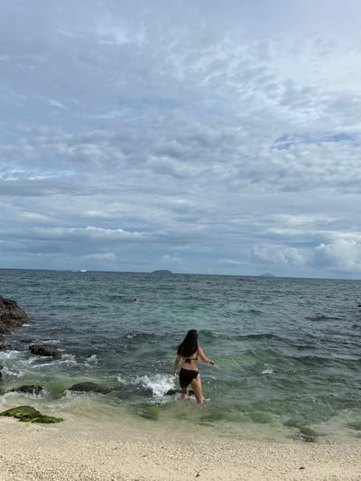 A woman in a brown bikini walks into the shallow, greenish-blue water of a beach in Phru Nai, Thailand. The sand is light-colored and composed of small pebbles and shells. Rocks covered in algae are visible in the water near the shore, and waves are breaking around them. In the distance, the ocean extends to the horizon, under a sky filled with a mix of gray and white clouds. A few small islands are visible on the horizon. A boat is seen in the distance on the left side of the image, and another person is swimming in the ocean further out. The overall scene is overcast and the water appears somewhat rough.