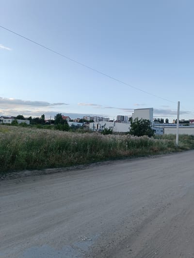 This image captures a view of a rural or semi-urban landscape in Durlești, Moldova. In the foreground, a dirt road runs diagonally across the frame, showing tire tracks and loose gravel. To the left of the road, a field of dry, tall grass and wildflowers extends towards a line of trees and buildings in the background. The buildings appear to be a mix of residential houses with red roofs and modern industrial or commercial structures, some of which are multi-story. The sky above is a pale blue with scattered, wispy clouds. A thin wire stretches across the upper portion of the image, suggesting the presence of power lines or communication cables. A solitary utility pole stands on the right side of the frame. The scene appears to be set during the late afternoon or early evening, given the soft, diffused lighting. There are no people or animals visible in the image.