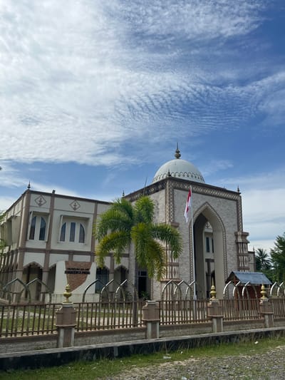 The image shows the exterior of the Museum Islam Samudra Pasai, located in the city of Kruengmate, Indonesia. The building features a dominant white dome and is constructed with light-colored stone and beige decorative elements. An Indonesian flag, red and white, is prominently displayed on a flagpole in front of the building, near the entrance. Palm trees are visible on the left and right sides of the building. A brown wooden fence surrounds the property, with decorative metal arches incorporated into its design. The sky above is blue with scattered white, wispy clouds. The ground in front of the fence is covered with small stones and some patches of grass. A sign that reads "MUSEUM ISLAM SAMUDRA PASAI" is visible in front of the building.