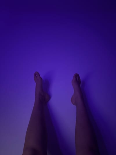 The image shows two bare legs and feet against a gradient of purple lighting, against a plain wall. The legs are positioned in the lower half of the frame, with the feet pointing upwards. The lighting creates subtle shadows. There are no other visible objects or people in the frame. The image does not contain information that allows for the determination of a specific location in Mönchengladbach, Germany.