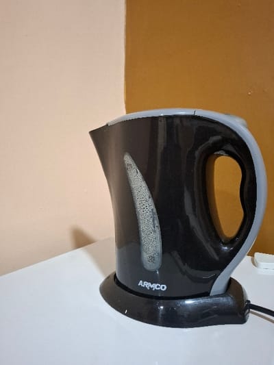 A black electric kettle with a gray handle and a black base sits on a white counter. The kettle has a visible water line and the words "ARMCO" on the base. The kettle is in a kitchen in Karagita, Kenya (latitude: -0.754, longitude: 36.437). The wall behind the kettle is painted a pale pink color on the left, and a darker yellow color on the right.  A white object is visible on the counter in the right background.  A black electrical cord is connected to the kettle, and it is plugged into a power outlet.  There is no one in the scene.