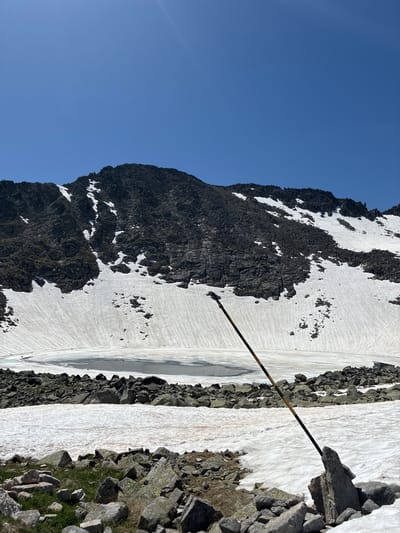 The image shows a mountainous landscape with snow-covered slopes and a frozen lake. A dark, rocky mountain peak dominates the background under a clear blue sky. In the foreground, there are scattered rocks and patches of snow. A black and yellow striped pole is visible, leaning diagonally across the scene, stuck in the rocky ground. This view is likely from the Rila Mountains near Samokov, Bulgaria, given the typical scenery of the region.