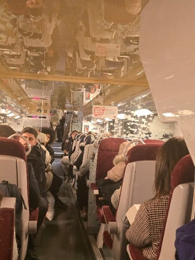 The image shows the interior of a train car filled with passengers. People are seated, with many wearing winter clothing. The seats are red and white. Overhead luggage racks are visible. There are cards or signs attached above some of the seats. The overall scene suggests a crowded train journey.