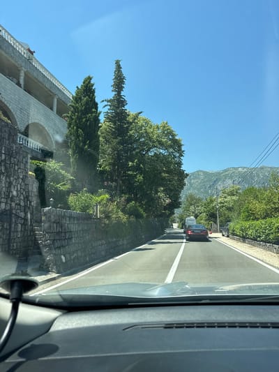 This is a vertical, low-angle shot taken from inside a vehicle, looking out at a road and a building. The scene is a scenic view of a mountainous landscape under a clear blue sky, suggesting a sunny day. The vehicle appears to be moving along a paved road with a white line marking the lane. A red car is visible ahead on the road, suggesting traffic. To the left of the road, there is a multi-story building with arched windows and a white railing along its balconies. The building is constructed with stone elements, and there are lush green trees and vegetation surrounding it. A stone wall lines the side of the road. The overall atmosphere is peaceful and summery. The time of day is likely midday due to the bright sunlight and clear sky. The location appears to be a coastal or mountainous region, consistent with the context of Perast, Montenegro, known for its stunning scenery. There are no people clearly visible in the image, but there are some objects like trees, cars, and parts of the vehicle the photo is taken from. The image captures a snapshot of a scenic drive.