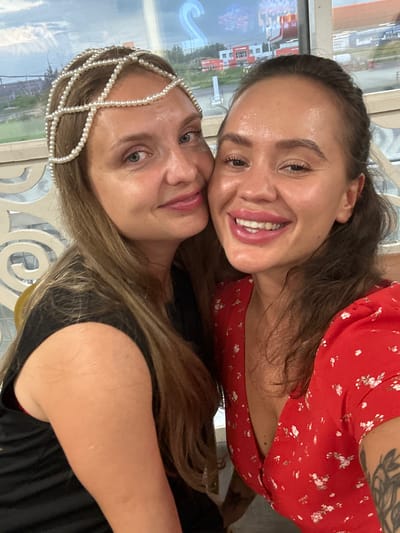 The media file is a vertical, close-up selfie of two young women, taken outdoors. The woman in the foreground is on the left, looking directly at the camera with a slight smile. She has blonde hair and is wearing a black top and a pearl headband. The woman behind her is on the right, smiling broadly with her mouth open, revealing her teeth. She has long brown hair and is wearing a red floral dress. Her left arm is visible, and it has a tattoo on it. The background appears to be a slightly blurred outdoor scene with some buildings and greenery, suggesting an urban or semi-urban environment. The lighting suggests it is daytime. The overall mood appears to be joyful and friendly. No visible text is present in the image.