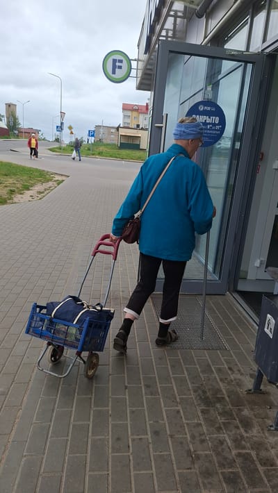 A person is walking towards a store entrance with a shopping cart. The person is wearing a blue jacket, black pants, and sandals. They have a bag slung over their shoulder. The shopping cart is blue and contains a large blue bag and other items. The store entrance has a glass door with a sign that says "FIX PRICE" and "ЗАКРЫТО" (Closed) in Russian. The surrounding area is a paved sidewalk with buildings and trees in the background. The sky is overcast. The scene appears to be in a city. The sign on the building indicates the location is a Fix Price store. Based on the presence of the Fix Price store and the overall urban environment, this image is likely located in a city within Belarus. The presence of Cyrillic text on the sign further confirms a Belarusian or Russian-speaking region.