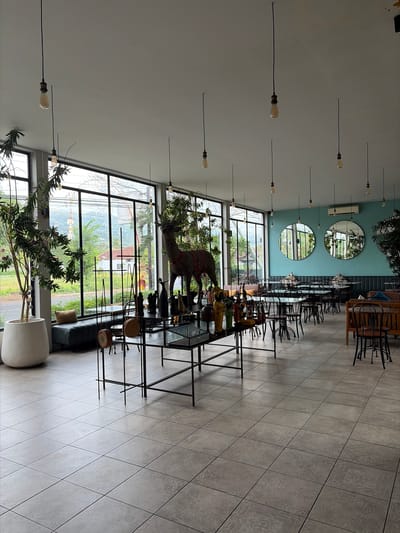 This image depicts the interior of a restaurant or cafe located in Banjar, Indonesia, characterized by its modern and airy design. Large glass windows dominate the space, offering a view of lush green hills and the surrounding landscape, suggesting a location on the outskirts of the city. Inside, the seating area is arranged with numerous tables and chairs, designed for a dining experience. Various decorative elements, including a large deer sculpture, ornamental statues, and potted plants, are strategically placed throughout the room, contributing to the establishment's aesthetic. The lighting fixtures, which consist of exposed bulbs hanging from the ceiling, add to the contemporary ambiance. The floor is tiled with large, light-colored tiles. In the background, two large circular mirrors are mounted on a teal-colored wall, reflecting the interior. The overall impression is one of a spacious and well-lit venue designed for comfort and visual appeal.