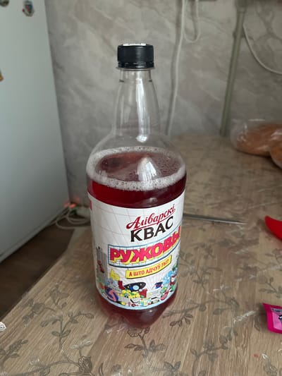 A partially filled plastic bottle of red kvass sits on a wooden table with a floral pattern. The label on the bottle features Cyrillic text and cartoonish illustrations. The background shows a light-colored wall with some electrical cords and a hint of a refrigerator. The scene is indoors, and based on the label's text which includes "Навагрудскі" (Navahrudski), suggesting a connection to the city of Navahrudak, Belarus, the image was likely taken in that location. There are no people or animals present in the image.