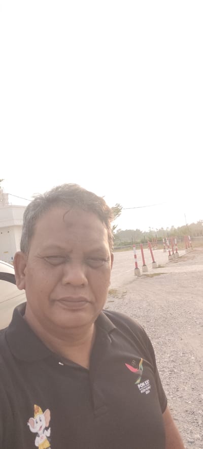 A medium shot selfie is taken from a slightly low angle, featuring a man in his late 40s or 50s wearing a black polo shirt. The man has brown skin, graying black hair, and is looking directly at the camera with his eyes partially closed. He has a neutral expression. The polo shirt has two emblems. On the left side of the chest, there is a cartoon of a white elephant wearing a yellow hat and yellow shorts, with Olympic rings on its shorts. On the right side of the chest, there is a logo with text that reads "PON XX" above "SUMATERA UTARA ACEH-SUMUT 2024". The man is standing outdoors in what appears to be a paved area with loose gravel. In the background, there are several red and white striped bollards arranged in a line. Further in the distance, there are trees and a clear sky with bright sunlight. The overall lighting suggests it is either sunrise or sunset. The location is identified as Meunasahaluedrienlhoksukon Barat, Indonesia.