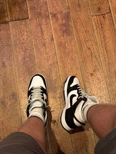 An overhead view captures the lower legs and feet of an individual in an indoor setting within Sigglesthorne, United Kingdom. The person is wearing dark, possibly charcoal-colored shorts, white ankle-length socks, and black and white high-top athletic shoes, specifically Nike Dunks, with light grey laces. The feet are positioned on a medium-brown wooden plank floor, which displays visible grain, minor scuffs, and dark speckling. A small, irregular brown liquid spill is present on the wooden floor near the right foot. Shadows are cast by the legs and shoes onto the floor.