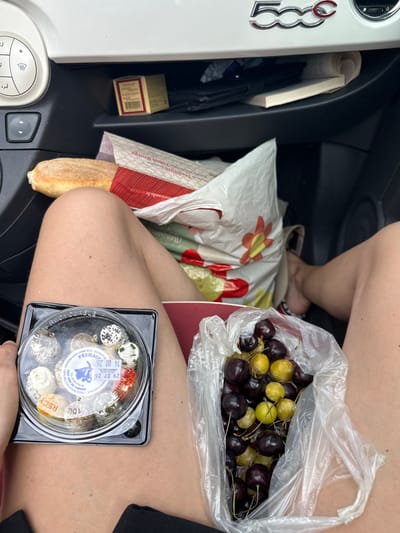 In Ouville-la-Rivière, France, a person is seated in the driver's seat of a Fiat 500 car. The person's legs are bare and visible in the foreground. On their lap, there is a clear plastic container filled with small, round cheeses. To the right of the cheese, a transparent plastic bag holds a collection of dark red and yellow cherries. Behind the person, a white shopping bag with a floral design is placed on the passenger seat. The car's dashboard is visible, featuring climate control dials and a "500" logo. A small brown box is tucked into a compartment on the dashboard, and a rolled-up book is also present. The overall scene suggests a pause during a journey, with the person enjoying snacks within the vehicle.