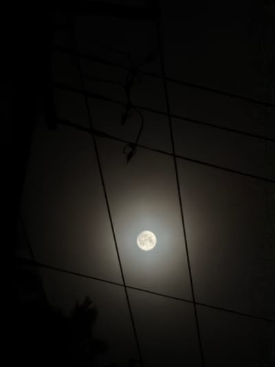 A full moon is visible in the night sky, obscured by power lines. The moon is a bright, circular object with visible texture on its surface. The surrounding sky is dark, with a hazy glow around the moon. Several dark, thin lines, presumably power lines, crisscross the frame horizontally and diagonally. In the lower left portion of the image, a dark, shadowy silhouette of a person's head and outstretched hand is visible against the brighter sky. The scene appears to be an outdoor urban or suburban environment. The location is Hampton Bays, United States of America.