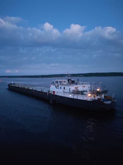 The image depicts a barge navigating a body of water, likely near Shelomovo, Russia, as the location was requested. The barge is long and dark, possibly carrying cargo, with a white and gray tugboat attached at the stern. The tugboat has lit windows and illuminated areas, suggesting operation in low light conditions. The water is dark and reflective, with subtle ripples indicating movement. A shoreline with trees is visible in the background. The sky is overcast with a mix of blue and gray clouds. The overall scene suggests twilight or dusk.