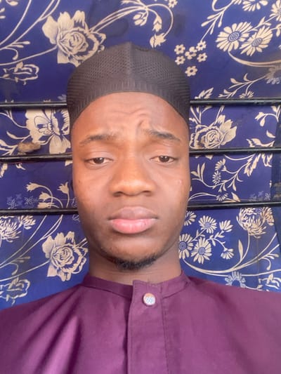 A young adult male is depicted in a close-up, head-and-shoulders portrait shot within an indoor setting in Katcha, Nigeria. He wears a black, mesh-textured kufi cap and a deep purple collared garment with a single circular, silver-toned decorative button visible at the neck closure. His skin tone is dark. His eyebrows are slightly furrowed, his eyes are open and directed forward, and his mouth is closed in a neutral expression. A faint beard and mustache are present. The background consists of a blue fabric featuring a repetitive pattern of light-colored floral and leaf motifs. Two horizontal dark metal bars are visible across the background, and a small, irregular brown object rests on the left side, positioned between the bars.