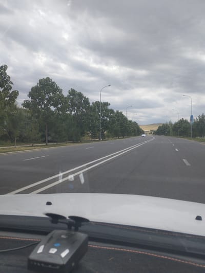 The image depicts a view from inside a vehicle traveling on a multi-lane road. The road is paved and has white lane markings. On either side of the road, there are rows of trees. Streetlights are visible along the roadside. In the distance, there is a white car traveling in the same direction. The sky is overcast with clouds. The foreground shows the dashboard of the vehicle with a radar detector mounted on it. The location is Birlashgan, Uzbekistan.
